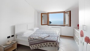 Ein Schlafzimmer mit Bett, Nachttisch, Kommode, einem Fenster mit Meerblick und einer Heizung.