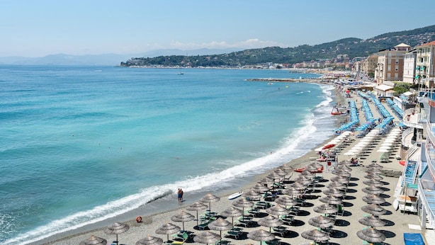Une plage avec des parasols et des chaises longues, une mer d’un bleu limpide et des bâtiments qui bordent le rivage.