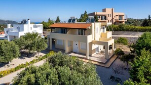Una casa moderna de dos pisos con techo plano, paneles solares y un jardín con olivos.
