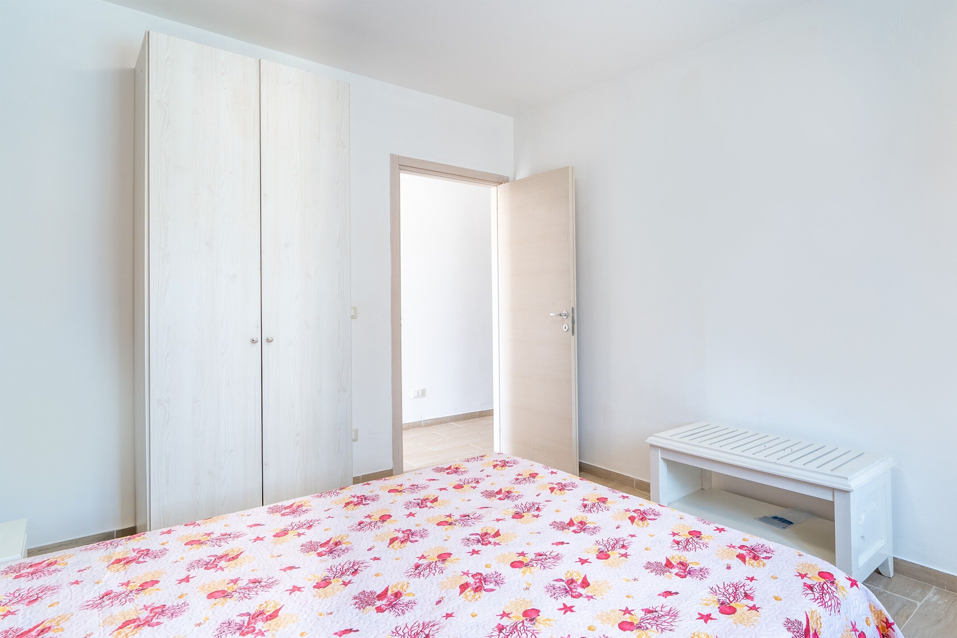A bedroom with a bed, a wardrobe, a bench, and an open door leading to a balcony.