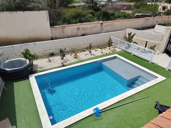 Une piscine rectangulaire bordée de blanc, entourée de gazon synthétique, d’une clôture blanche et de petites plantes.