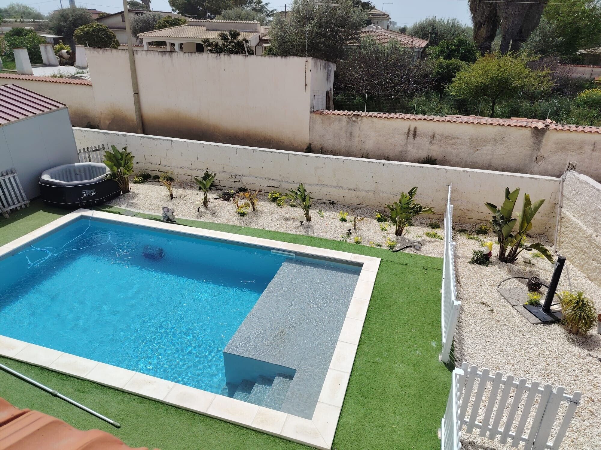A rectangular swimming pool with a tiled edge, surrounded by artificial grass and a gravel area with small plants.