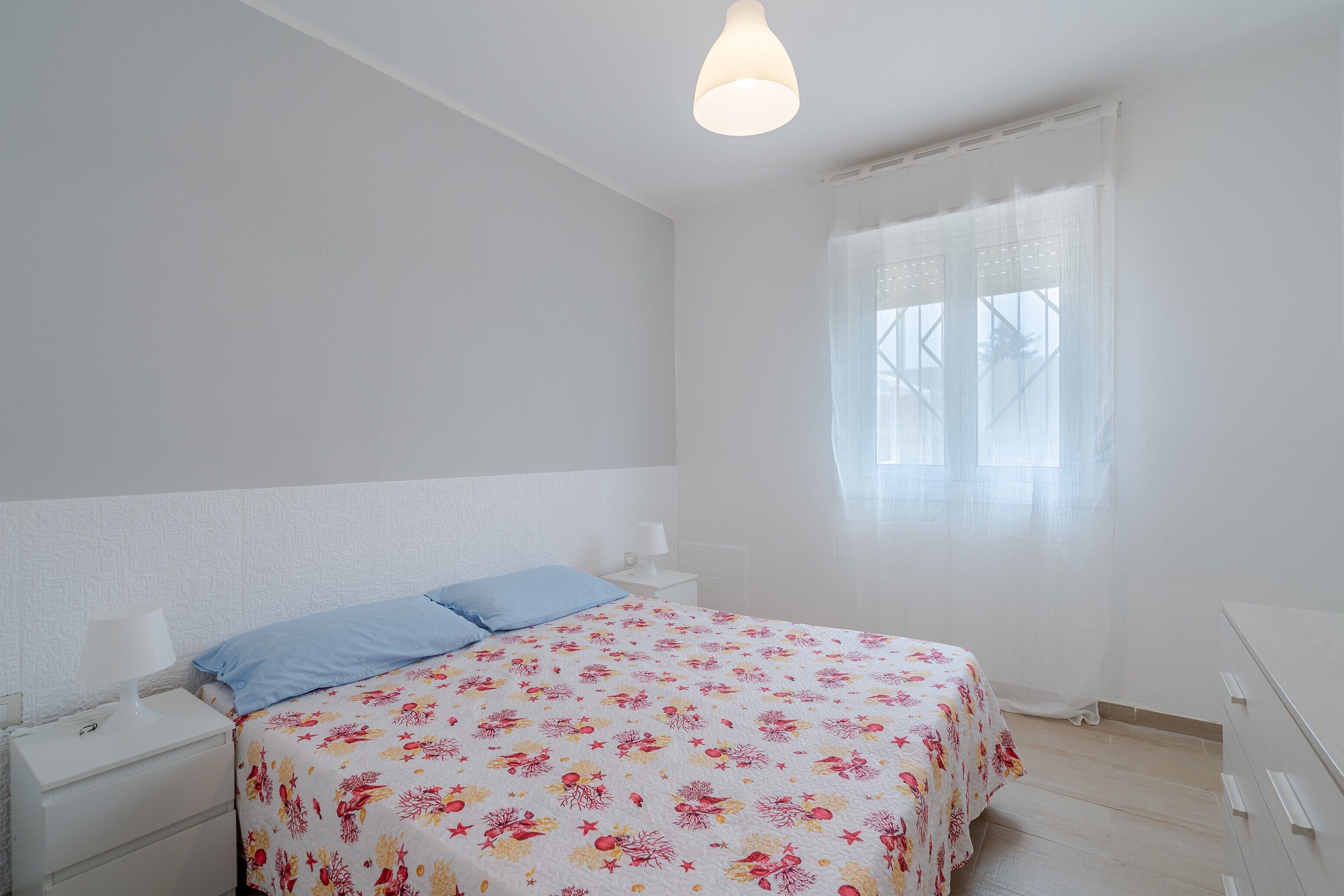 Ein Schlafzimmer mit Bett, Nachttisch, Kommode und einem Fenster mit durchscheinenden Vorhängen.