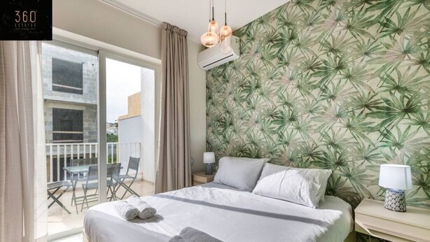 Hotelkamer met een bed, twee bedlampen, een raam met gordijnen, een balkon met een tafel en stoelen, en tropische behang.