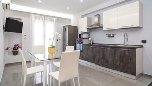 A modern kitchen with a glass dining table, white chairs, a refrigerator, microwave, oven, and a sink. - Apartment "Dharma" with Mountain View, Terrace & Wi-Fi (Giardini-Naxos)