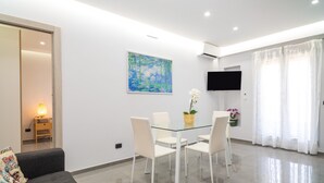 A modern living room with a glass dining table, white chairs, a sofa, a painting, a TV, and a window with curtains. - Apartment "Dharma" with Mountain View, Terrace & Wi-Fi (Giardini-Naxos)