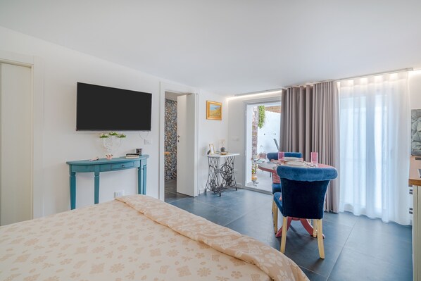 Un dormitorio con cama, televisor, escritorio, silla y zona de comedor con mesa y cortinas.