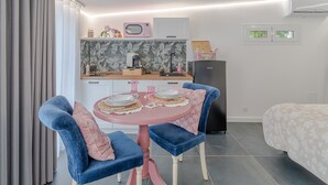 Una habitación compacta con kitchenette, un área de comedor con una mesa rosa y sillas azules, una cama con colcha estampada floral y una ventana con vista a vegetación.
