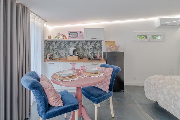 A compact room with a kitchenette, a dining area with a pink table and blue chairs, a bed with a floral bedspread, and a window with a view of greenery.