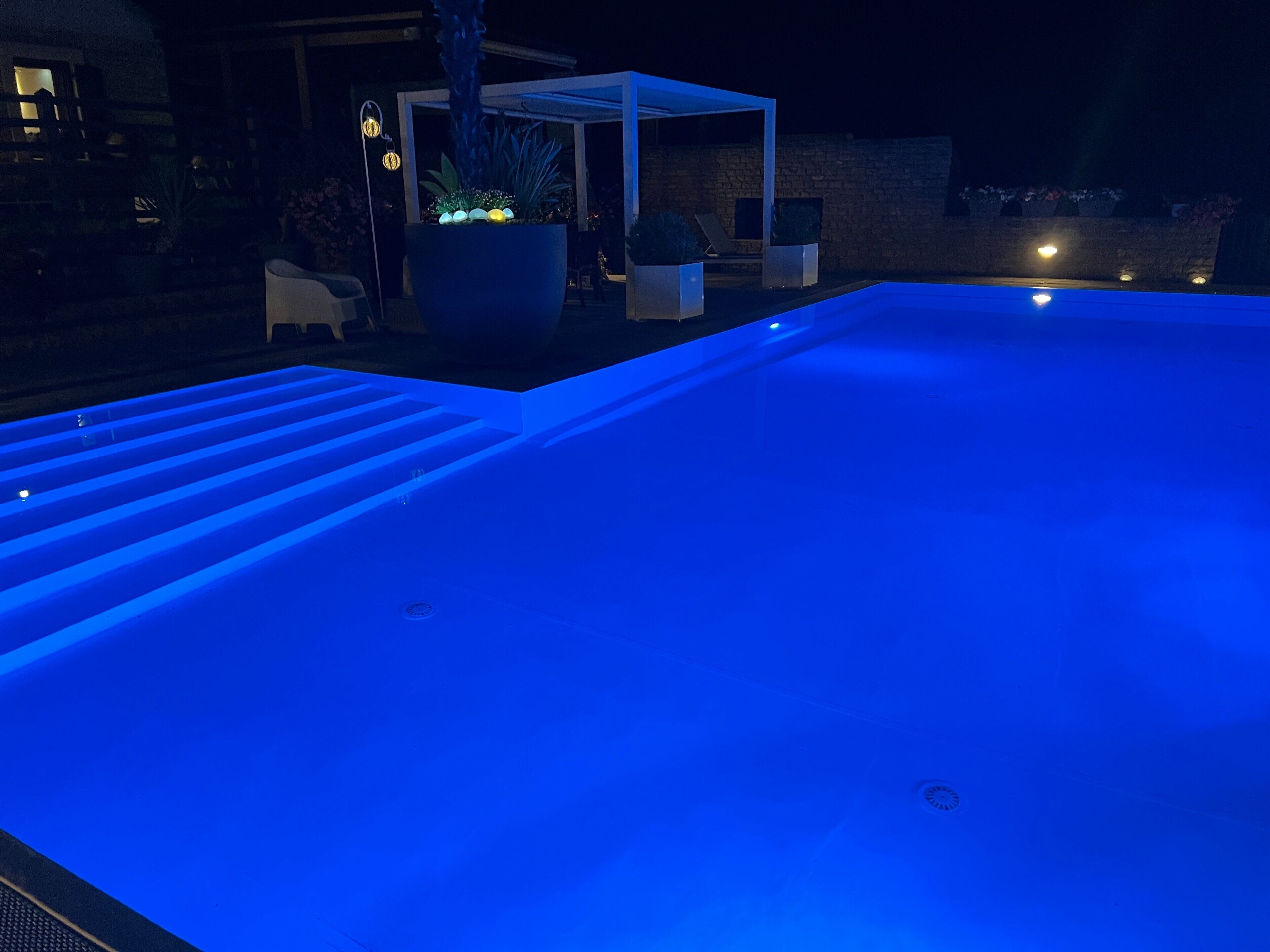 Ein Außenpool bei Nacht mit blauer Beleuchtung, einer überdachten Lounge und Topfpflanzen.