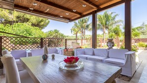 A covered patio area with a wooden table, white cushioned seating, and a decorative vase. - Apartment 'Giusa' with Shared Pool, Wi-Fi and Air Conditioning (Caltagirone)