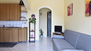 A living room with a grey sofa, a television, a kitchen area with wooden cabinets, and a hallway leading to a bathroom. - Holiday Apartment "Corbezzolo" with Balcony & Shared Terrace (Bari sardo)