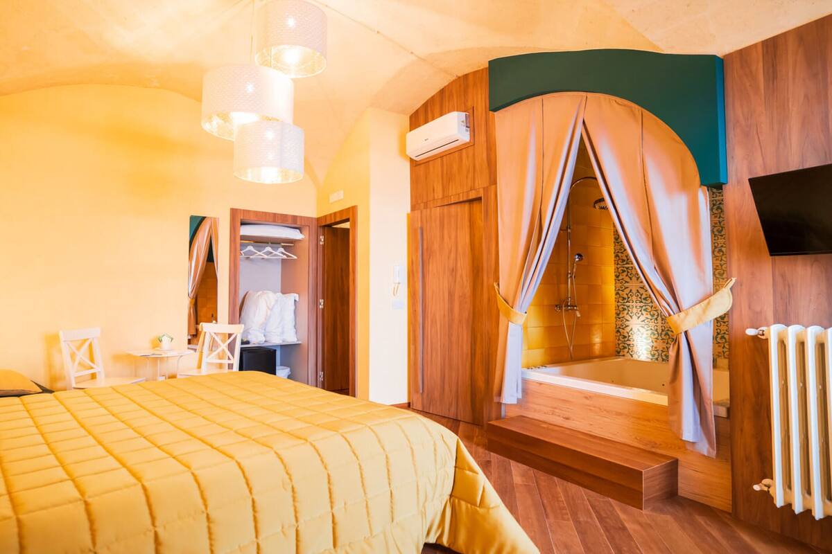 A bedroom with a bed, a small table, a chair, a wardrobe, an air conditioning unit, a shower area with curtains, and a television mounted on the wall.