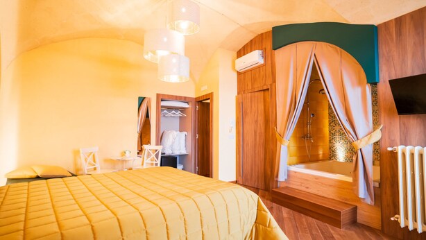 Quarto com cama, uma pequena mesa, cadeira, guarda-roupa, ar condicionado, área de chuveiro com cortinas e televisão na parede.