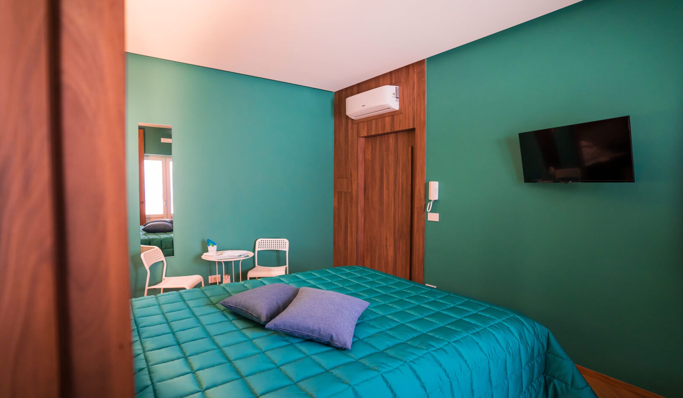 A room with teal walls, a bed with a teal quilt, a wooden door, a mounted TV, a small table with chairs, and a window with a view.