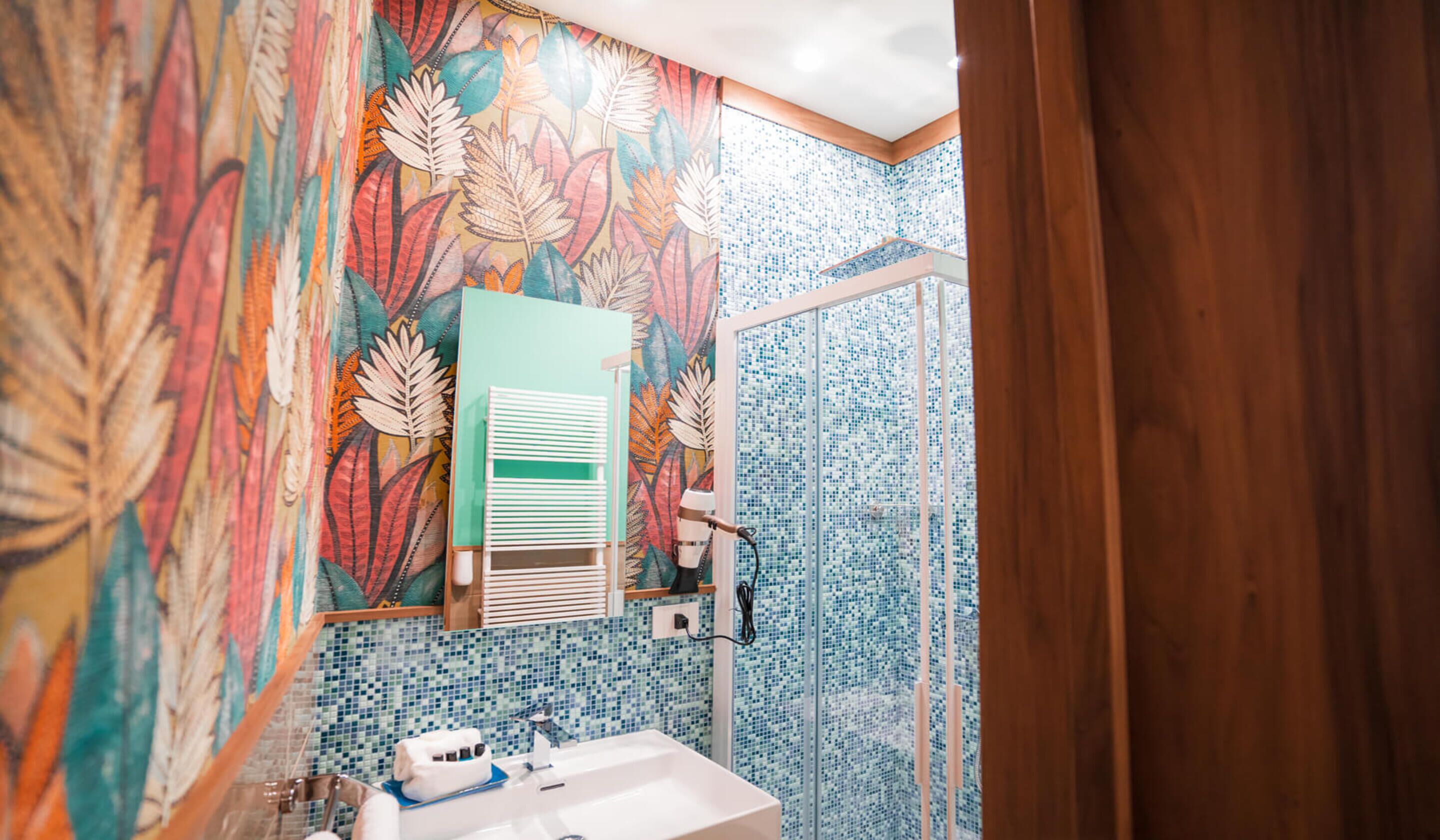 Une salle de bain avec un mur décoré d’un motif coloré représentant des feuilles, un lavabo blanc et un espace douche.