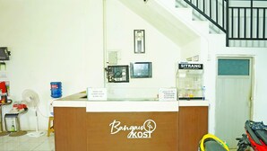 En receptionsdisk med et skilt der siger "Bangun KOSTI". Der er to skærme på væggen, en ventilator og en vanddispenser.