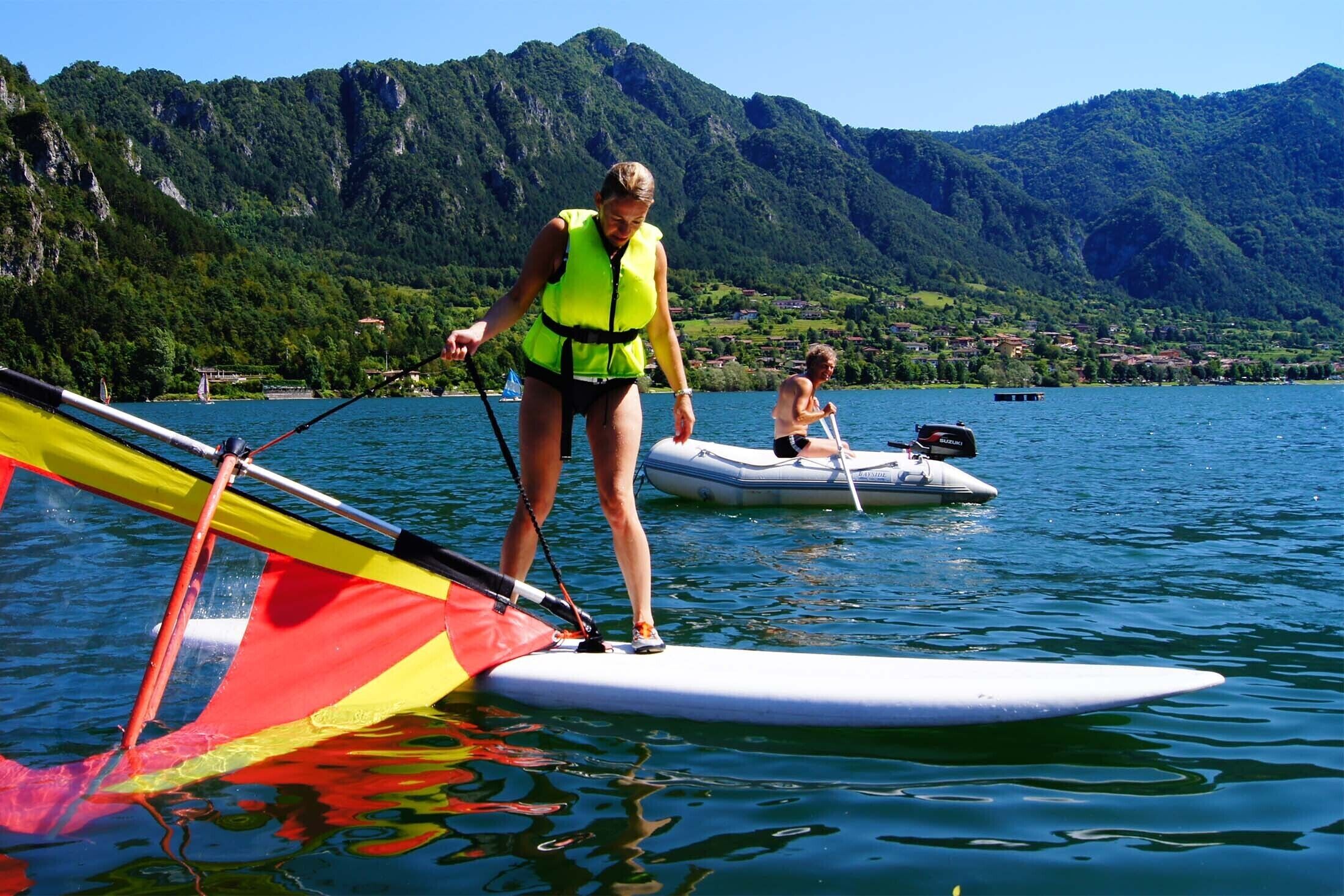 A person standing on a paddleboard with a sail, another person paddling a boat, and a mountainous landscape in the background.