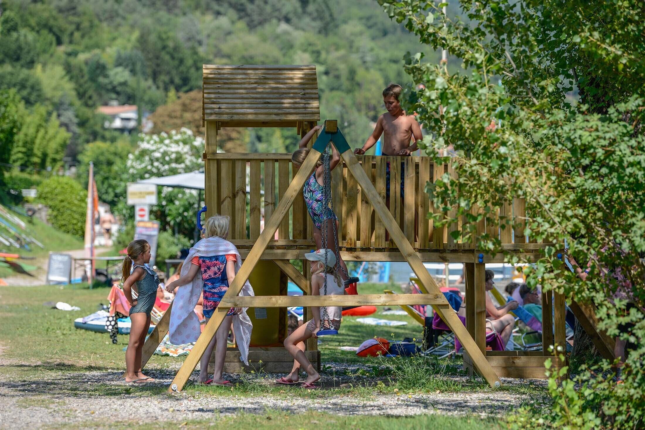 A wooden play structure with a slide and climbing area, surrounded by a grassy area with people.