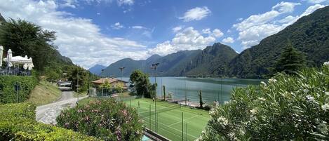 A tennis court with a mountain backdrop, surrounded by lush greenery and a clear sky.