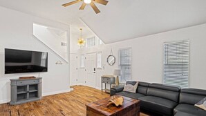 A modern living room with a wooden floor, a black leather sofa, a wooden coffee table, a TV, and a staircase leading to an upper level. - Modern Comfort: WiFi, 3BR, King, Patio (Round Rock)