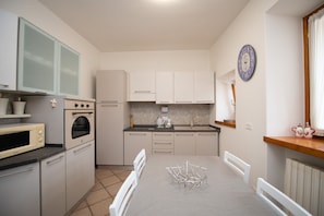 Una cocina compacta con armarios blancos, un microondas, un horno y un lavamanos. Hay una mesa de comedor con sillas y un reloj en la pared.