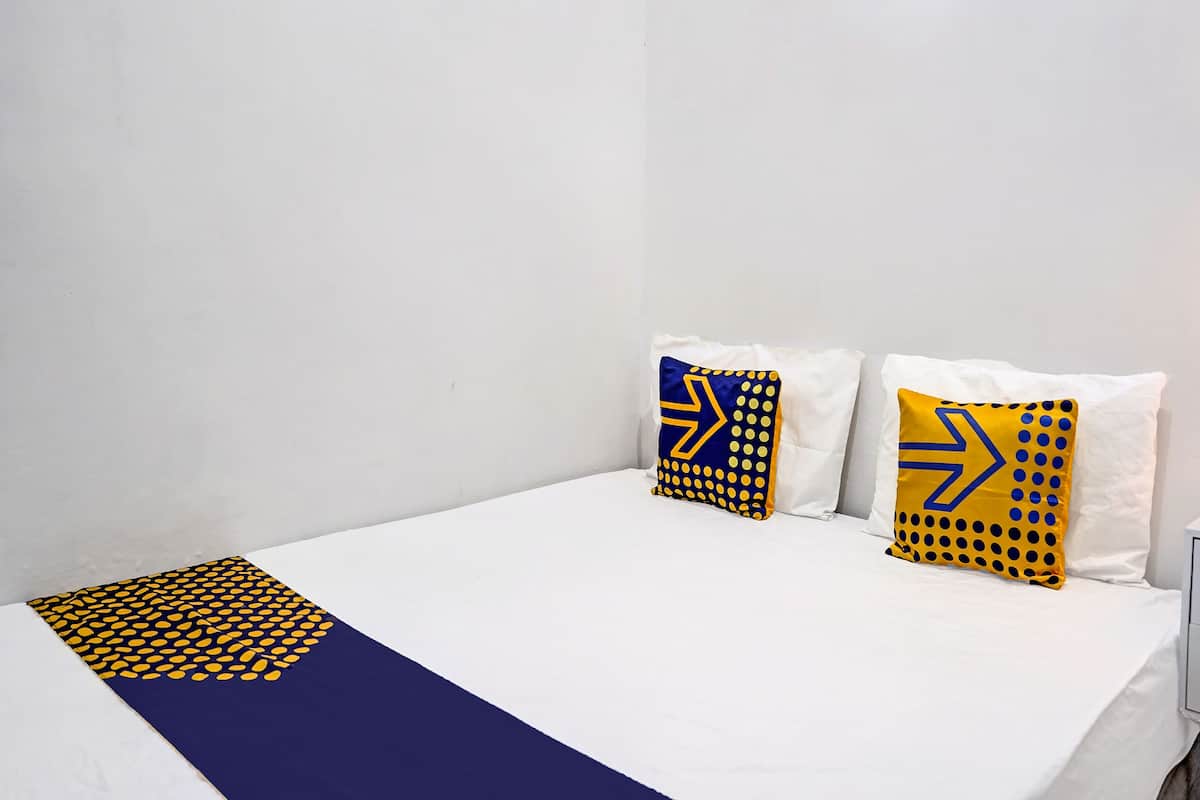 A small, single-bed room with white walls and a white bedspread. There are two pillows with yellow and blue patterns on the bed.