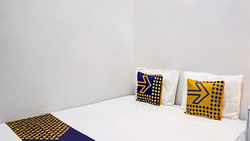 Une petite chambre avec un lit simple, aux murs et au couvre-lit blancs. On trouve deux oreillers à motifs jaunes et bleus sur le lit.