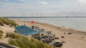 A beach with wind turbines in the background, a lifeguard station, and a sandy beach area with people and vehicles. - Holiday Home near Beach Burgh-Haamstede (Burgh Haamstede)