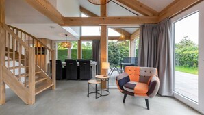 A spacious room with a wooden staircase, a dining area with chairs and a table, a colorful armchair, and large windows offering a view of greenery. - Holiday Home near Beach Burgh-Haamstede (Burgh Haamstede)
