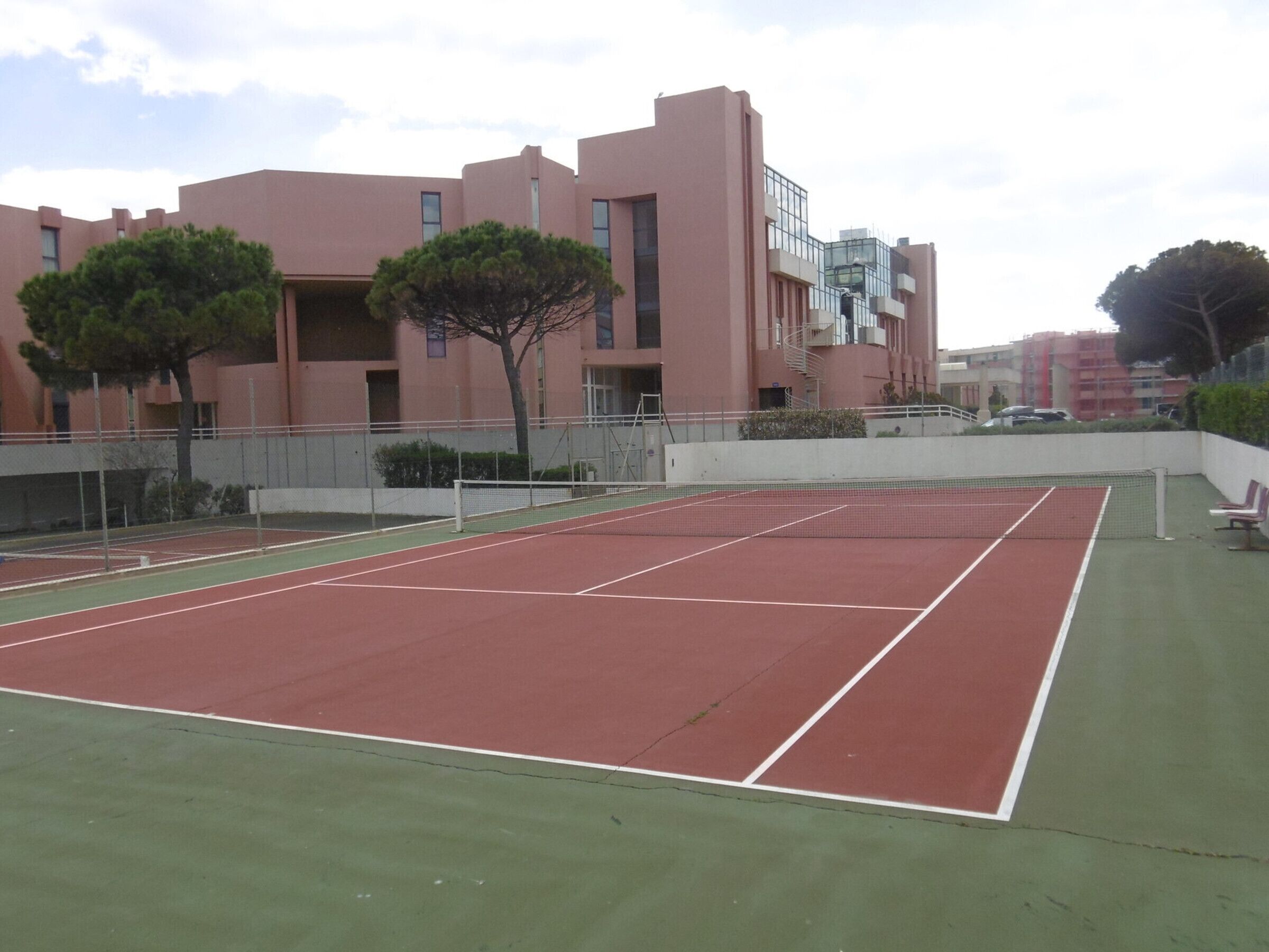 An outdoor tennis court with a net, surrounded by a low fence and trees.
