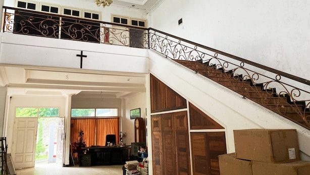 Un intérieur spacieux avec un escalier, une croix au mur et un bureau sur lequel se trouvent divers objets.
