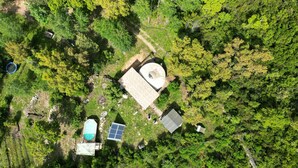 An aerial view of a property with a swimming pool, solar panels, a circular building, and a rectangular building surrounded by dense forest.