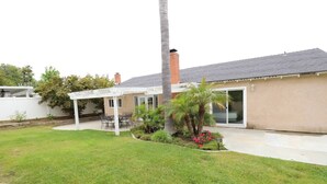 Una casa con porche cubierto, césped y un pequeño jardín con palmeras y flores.
