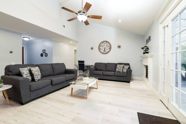 A modern living room with two grey sofas, a glass coffee table, and a ceiling fan. - Extended Stay Oasis w/ yard & Arcade (Maple Perch) (Mission Viejo)