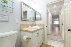 Une salle de bain moderne avec des toilettes, un lavabo et des miroirs. Un panneau indiquant l’emplacement d’une poubelle est accroché au mur, et une plante est posée sur une étagère.