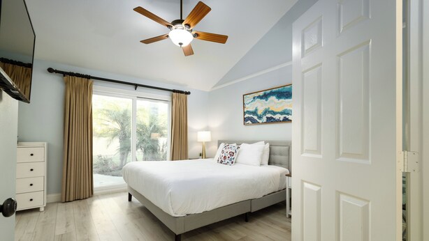 Un dormitorio con una cama, ventilador de techo, ventana con cortinas, un cuadro en la pared y una puerta.