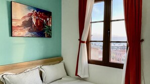 Una cama con ropa blanca, un cuadro en la pared y una ventana con cortinas rojas.