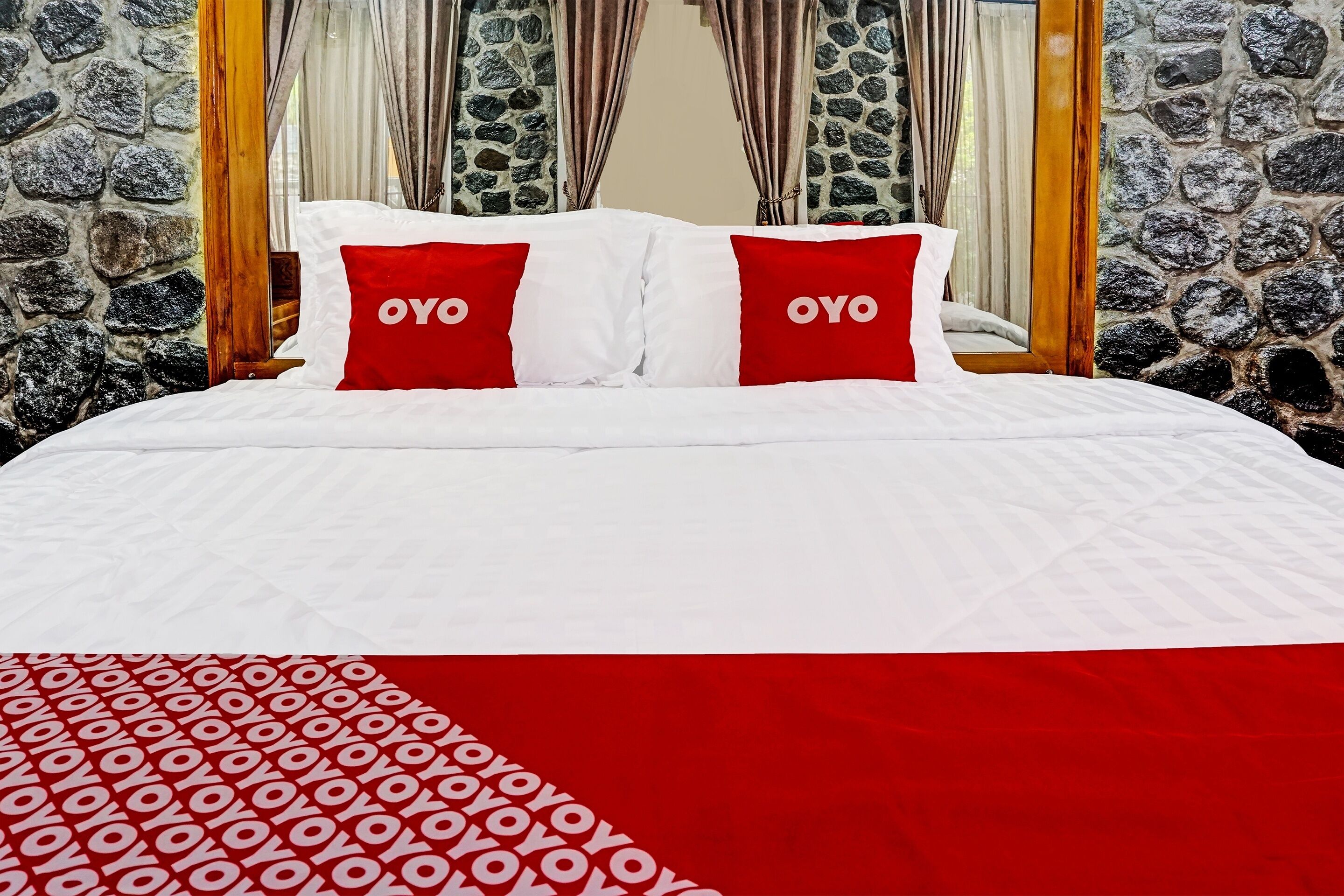 A bed with white and red bedding, two red pillows with the word "OYO" on them, and a stone wall behind.
