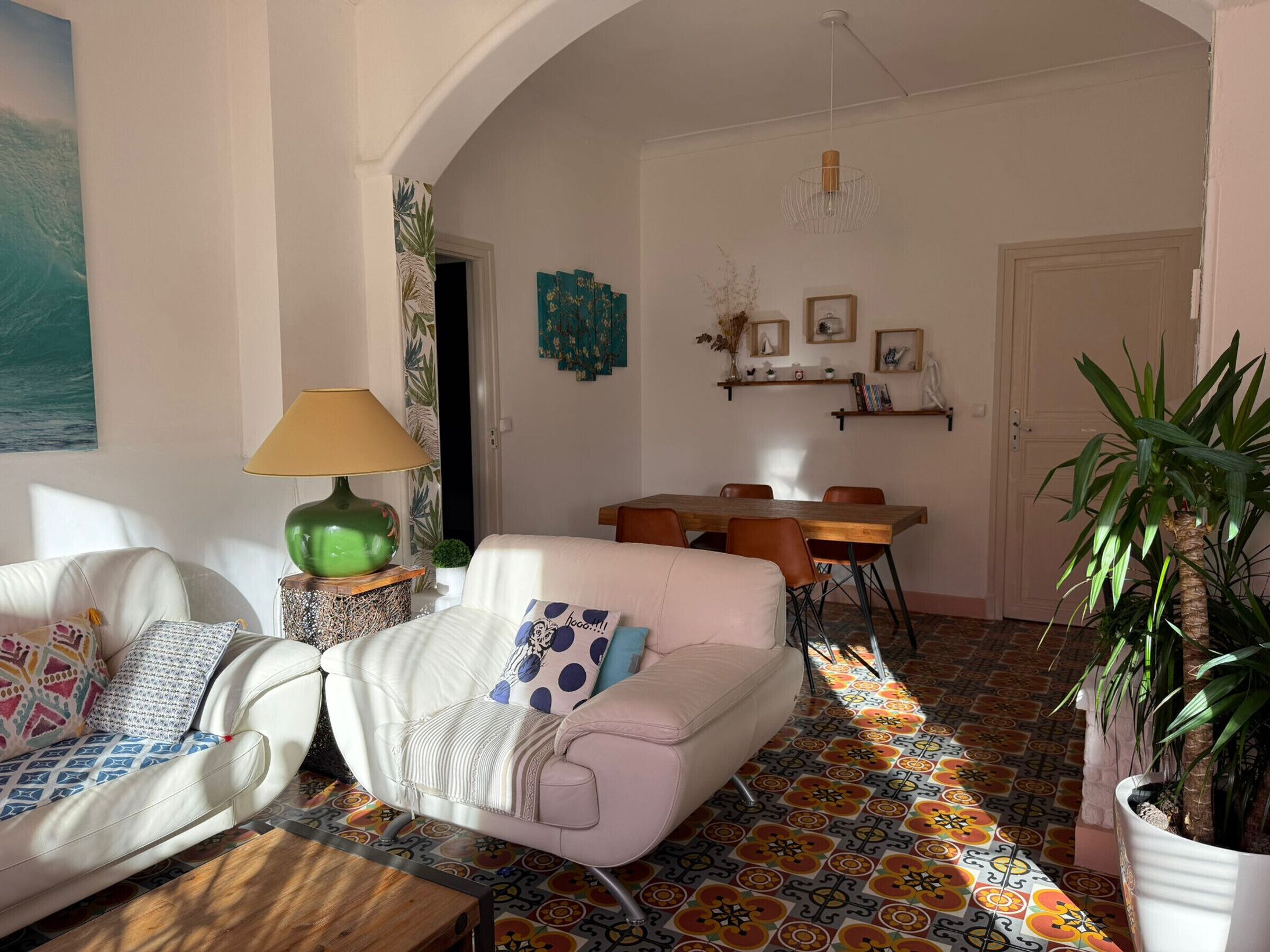 Uma sala de estar com um sofá branco, um abajur verde, uma mesa de jantar com cadeiras e um chão com ladrilhos coloridos.