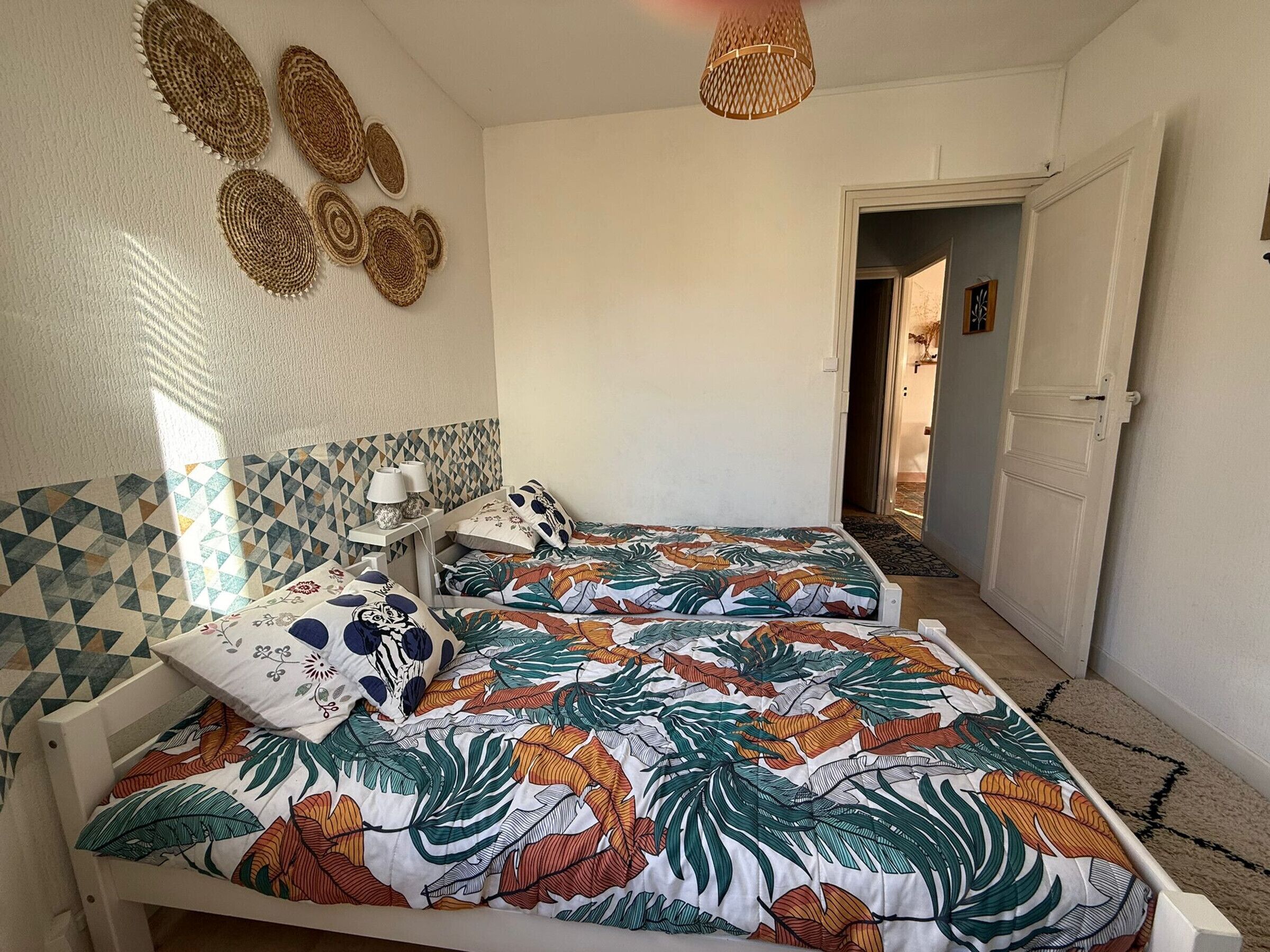 A bedroom with two beds, a geometric-patterned wall, a woven headboard, and a door leading to another room.