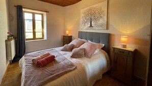 Un dormitorio con techo de madera, una cama con ropa blanca, dos lámparas de noche, un radiador, una ventana con cortinas y un cuadro de un árbol.