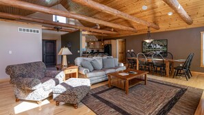 A spacious living room with a high wooden ceiling, a grey sofa, patterned armchairs, a wooden coffee table, and a dining area with a wooden table and chairs.