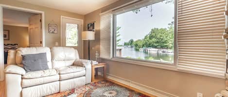 A living room with a beige sofa, a patterned rug, a side table with a lamp, and a large window with blinds overlooking a lake.