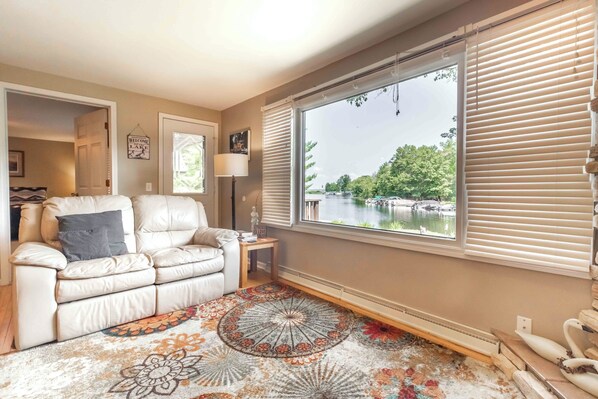 A living room with a beige sofa, a patterned rug, a side table with a lamp, and a large window with blinds overlooking a lake.
