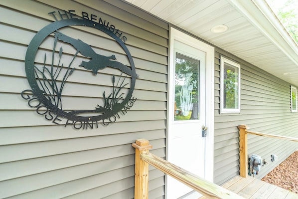 A metal sign with a bird and plants design, and the words "The Bench's Goose Point Cottages" on a house exterior.