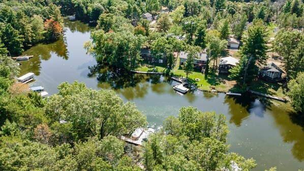 An aerial view of a lakeside property with multiple boats, a dock, and residential buildings surrounded by dense trees.