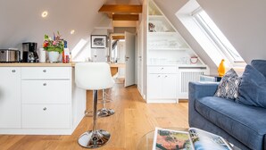 A modern loft-style apartment with a kitchen, dining area, and living room. It features a sloped ceiling with skylights, wooden beams, and white cabinetry. - Modern. Ruhig. Wiesenblick. Ringhoog Hüs Whg. 5 "weitblick" Objekt-id: Rhh/05 (Westerland)
