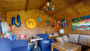 Interior de una cabaña de madera con un sofá azul, una mesa de madera, un escritorio pequeño y un cuadro de una escena tropical.