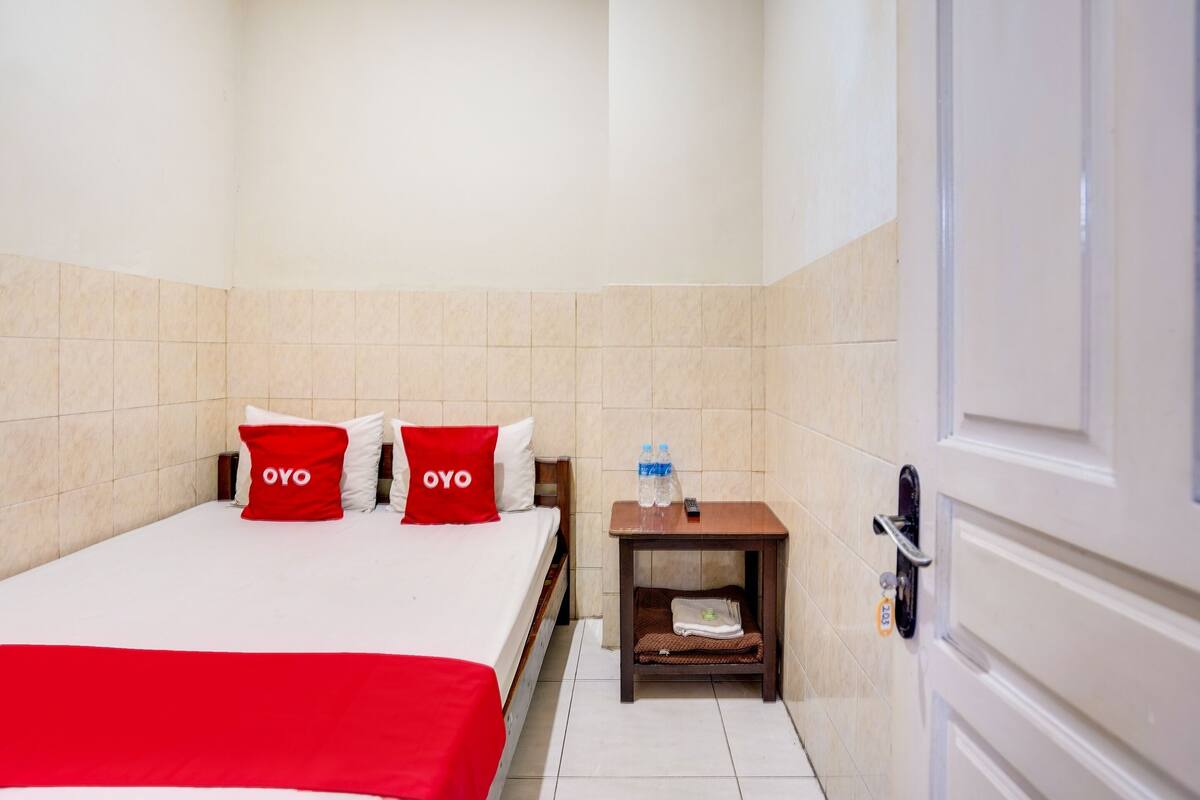 Een hotelkamer met een bed, twee rode kussens, een nachtkastje met een fles en een afstandsbediening, en een deur met een slot.